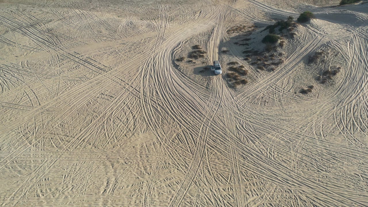 Aerial frame from Los Cabos, Mexico, showing a white car on pale desert sand beside clustered shrubs and crossing tire tracks, with no visible people.
