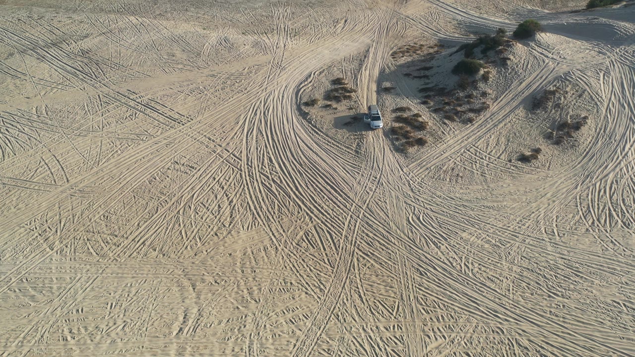 Aerial Los Cabos frame with the white car slightly nearer the center, a winding sandy road curving around low shrubs, and no visible people.
