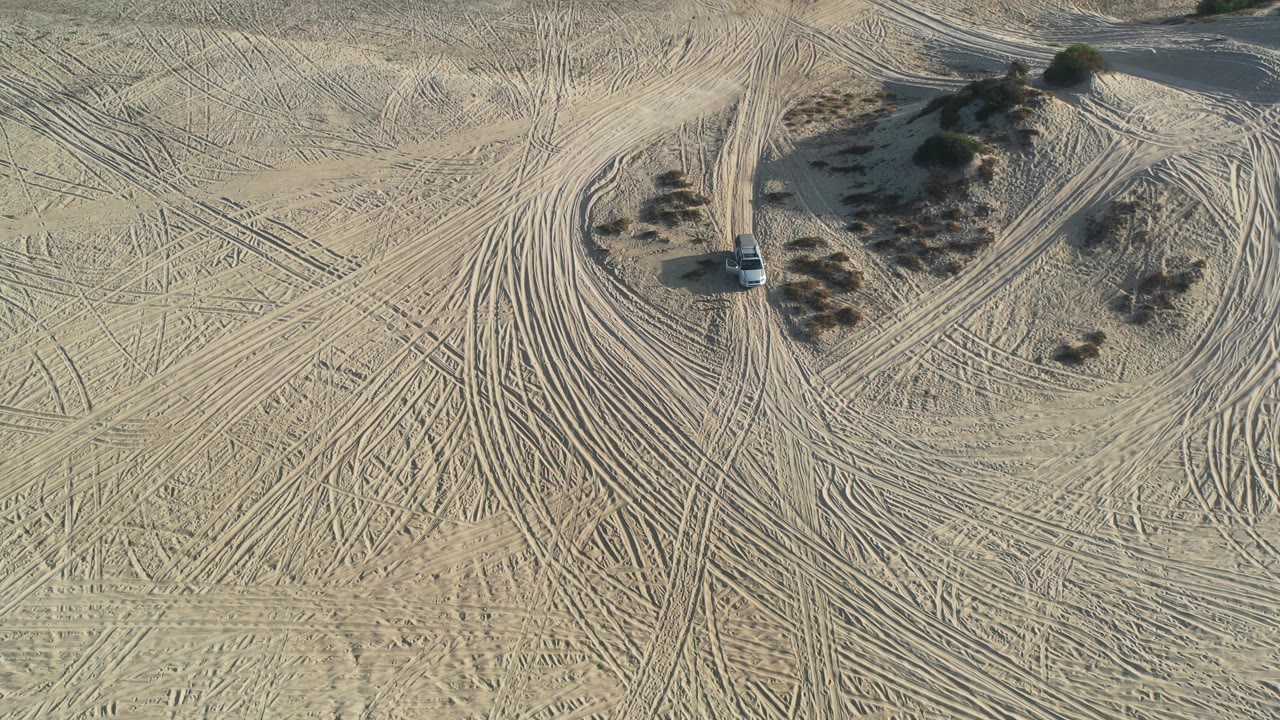 Overhead desert view with a car near scrub and multiple tire paths.