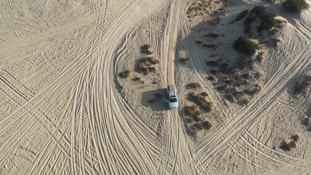 Overhead view in Los Cabos, Mexico of a white car on pale sand beside scrub patches and dense tire tracks, with no visible people.