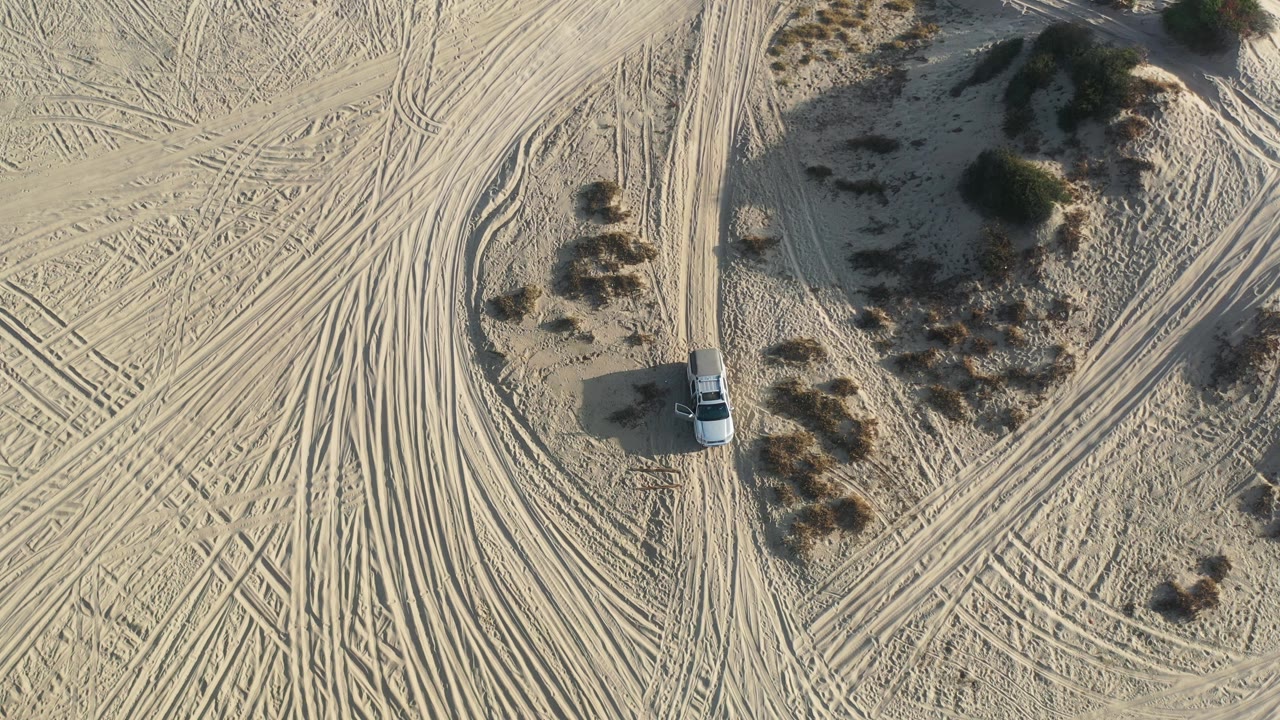 Overhead Los Cabos, Mexico frame with the white car near the lower center, tire ruts crossing the sand, and shrubs casting shadows, with no visible people.