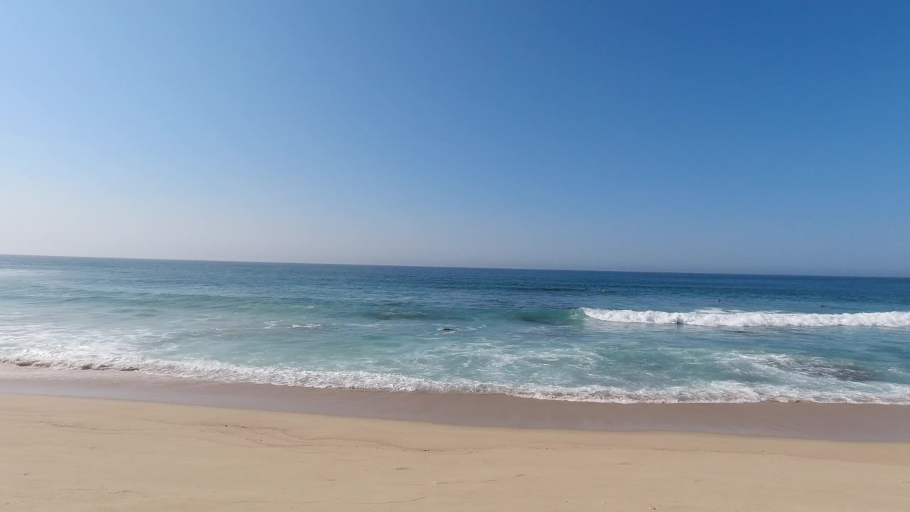 Los Cabos shoreline with a broad band of pale sand, white surf spreading across the beach, dark rocks near the waterline, and no visible people.