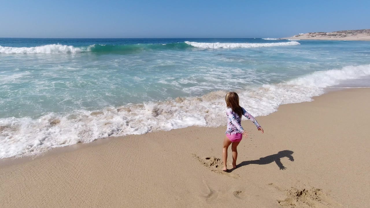 The child remains near the wet sand in Los Cabos while a low wave spreads toward her from the turquoise water.