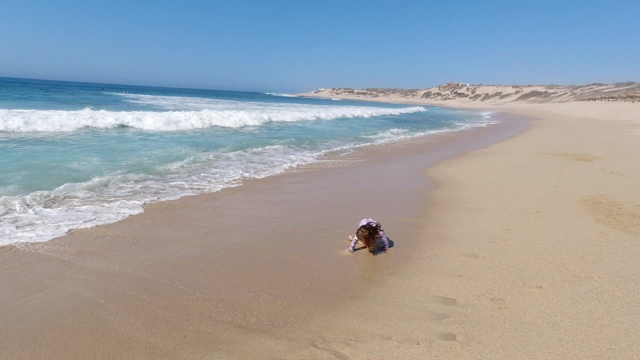 Young girl crouching on sandy beach near breaking waves.