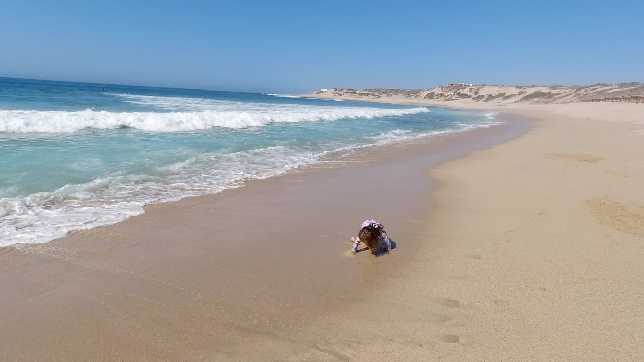 One second in, the girl remains crouched on the Los Cabos shoreline as a band of foamy water spreads across the left foreground.
