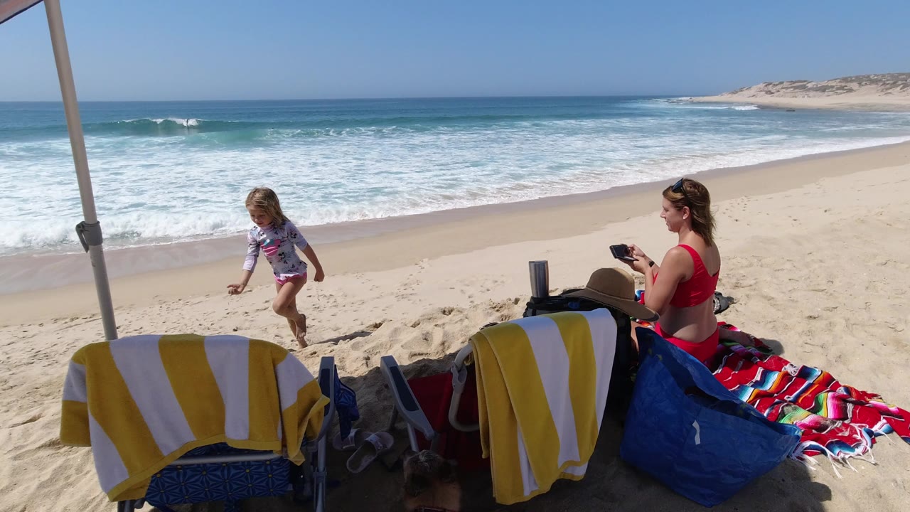 Woman sitting under a beach umbrella as a child walks on sand near the surf.