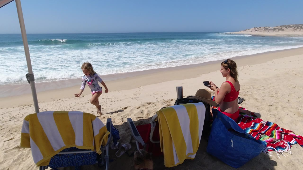 In Los Cabos, Mexico, the child is farther across the sand as the seated woman under the umbrella looks at a small dark object, with surf and dunes behind them.