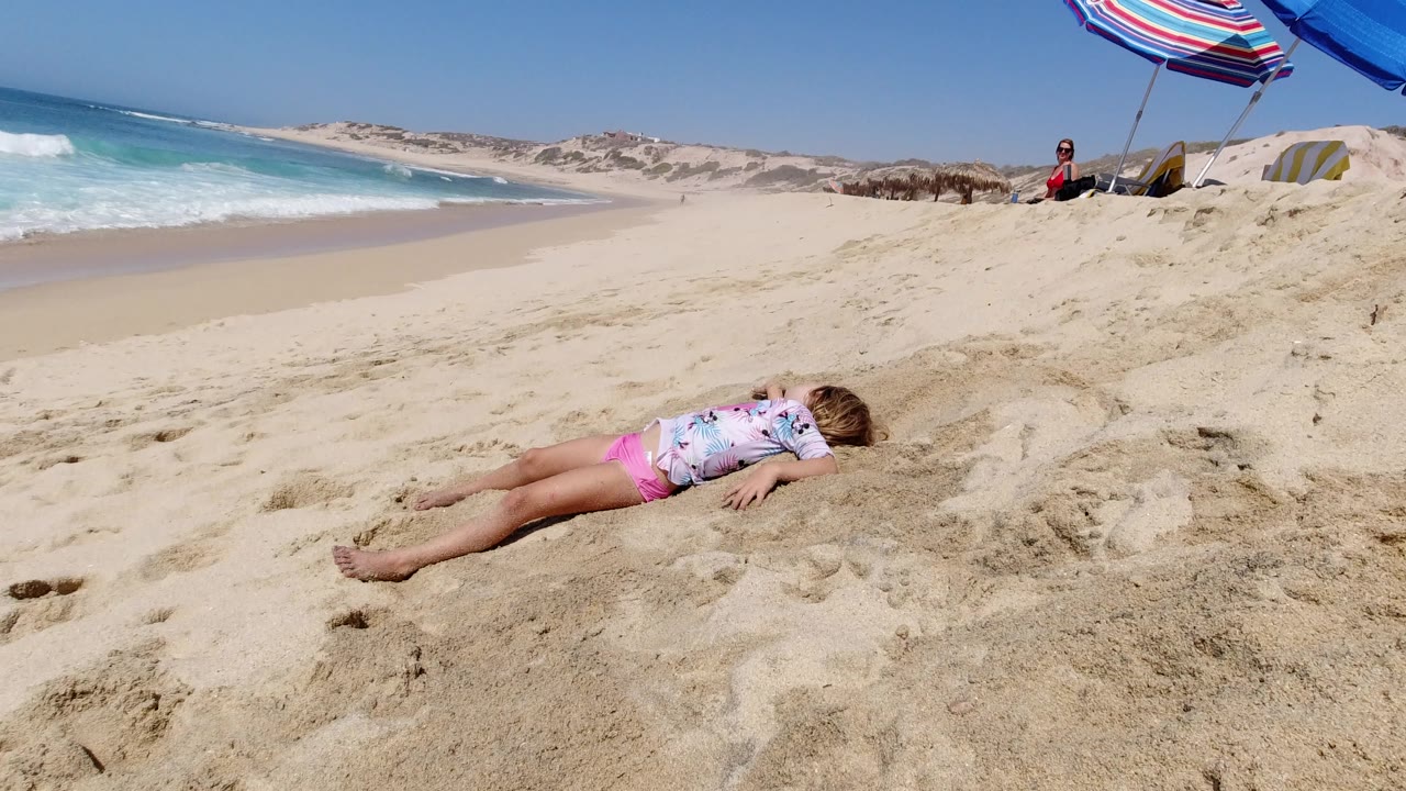 At 0:01, the child remains stretched out on the sand in Los Cabos, with the waterline curving behind them and a distant adult seated near beach shade.