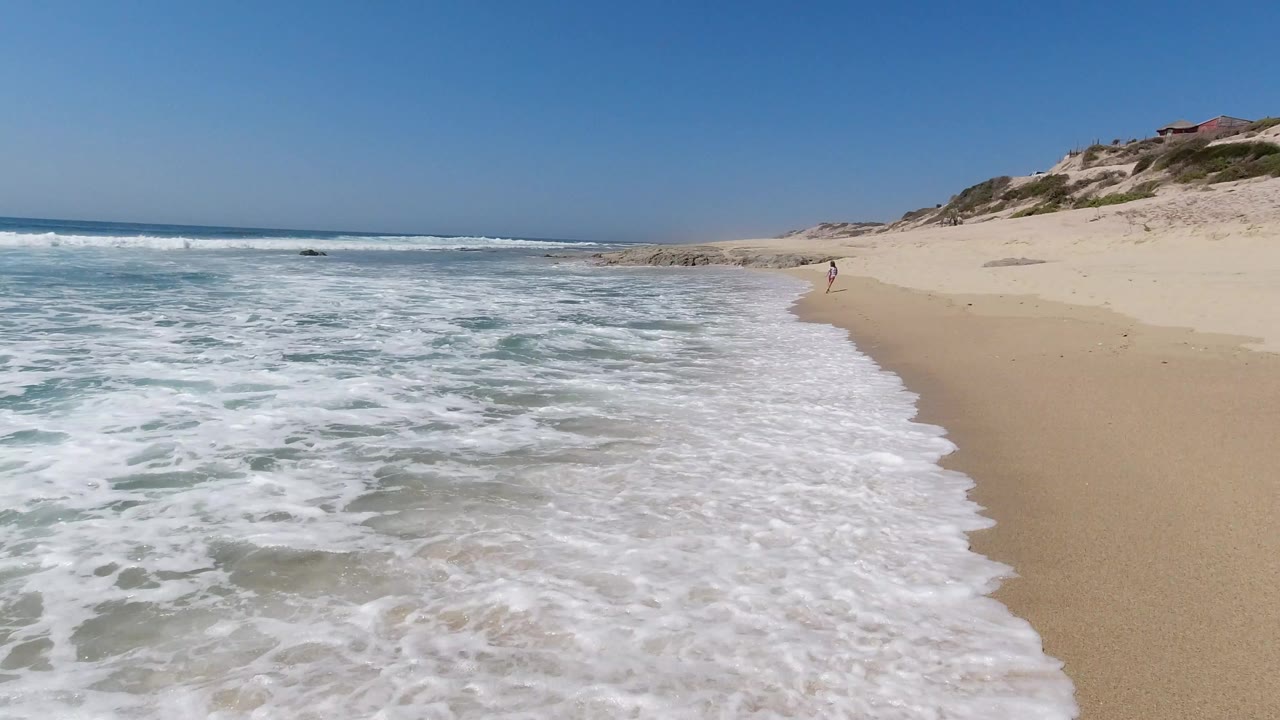 Opening frame of the Los Cabos shoreline with foamy water washing across wet sand, dunes to the right, and no recognizable people visible.