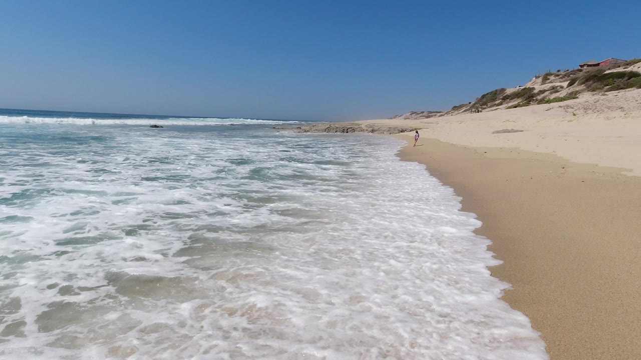 The Los Cabos beach remains framed wide as whitewater spreads farther across the foreground sand with dunes and scrub along the right side.