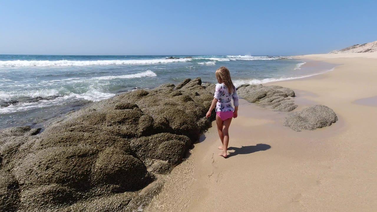 A young girl walks barefoot away from the camera on a Los Cabos beach, with dark rocks ahead, white surf to the left, and pale dunes in the distance.