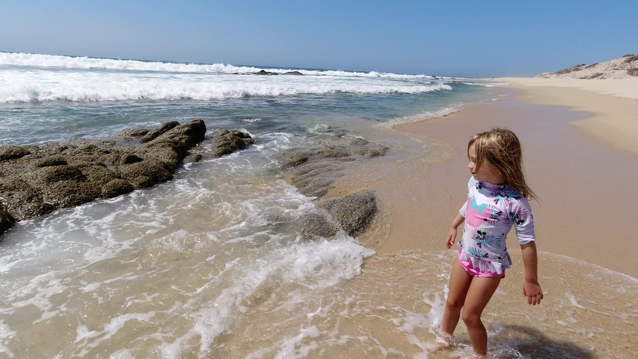The girl remains near the wet sand and rock edge while white foam spreads across the beach in Los Cabos.