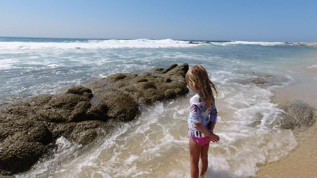 The child remains near the rocky Los Cabos shoreline while foamy water spreads across the sand and a line of surf breaks in the background.