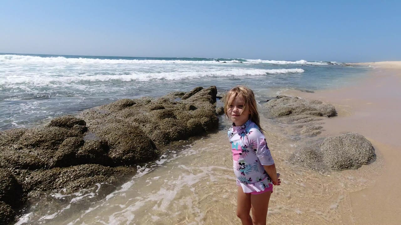 A young girl faces the camera on a Los Cabos beach, standing on wet sand beside dark rocks with foamy surf and blue sky behind her.
