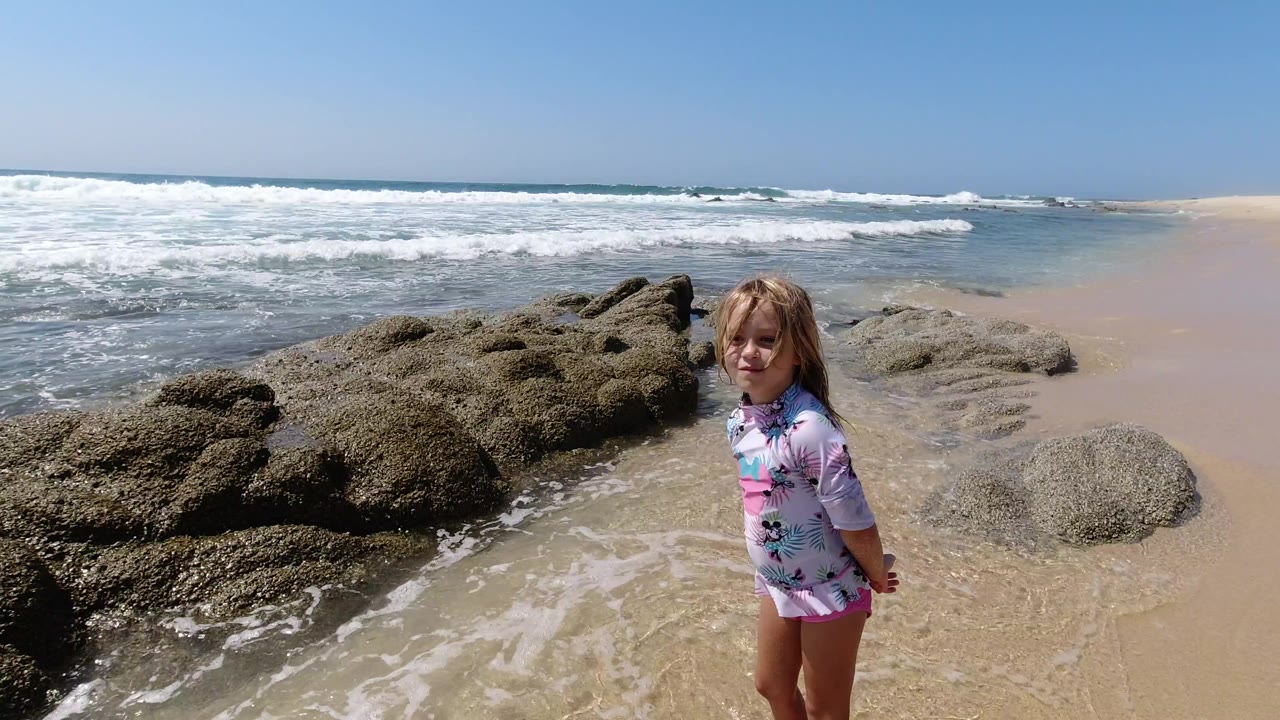 The girl stands near the center of the Los Cabos shoreline frame, with textured rocks to her left and whitewater spreading across the sand behind her.