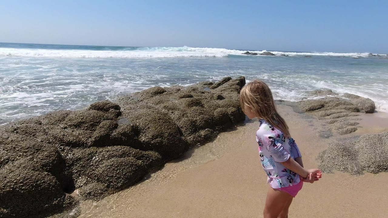 Young girl standing on sand beside dark rocks and ocean surf.