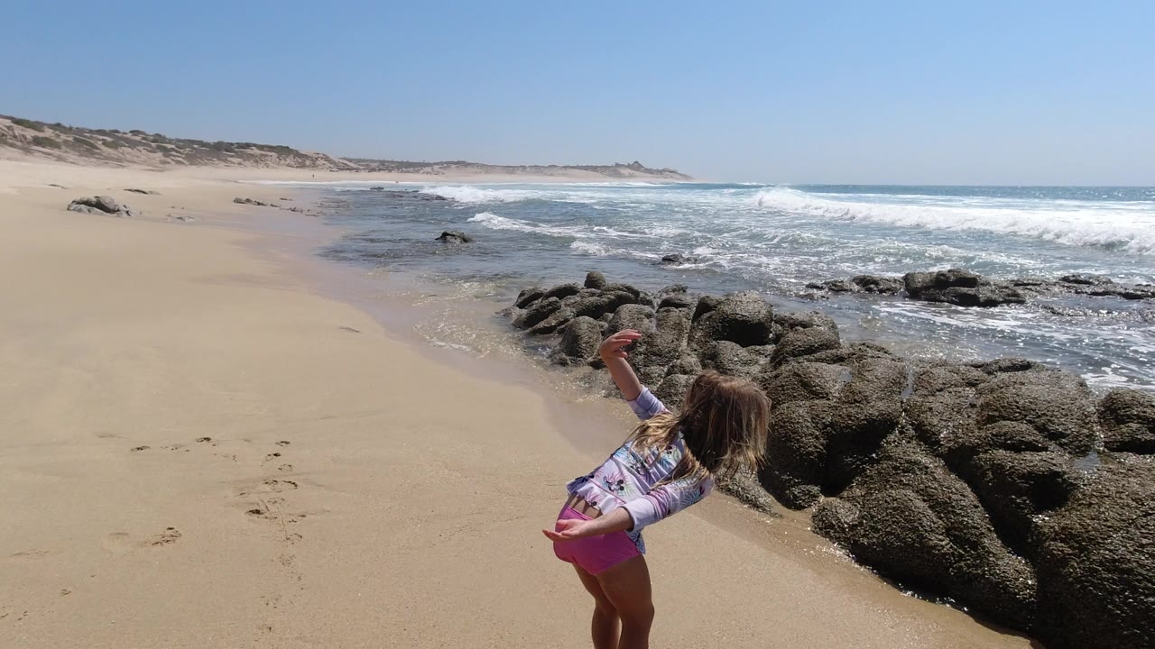 Young girl leaning toward rocks and surf on a sandy beach.