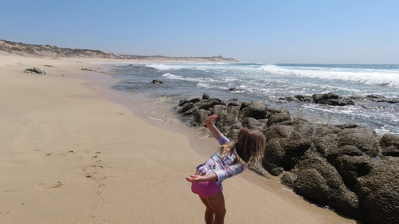 One second in, the girl reaches outward near the wet sand as ocean foam approaches the rocks on the Los Cabos shoreline.