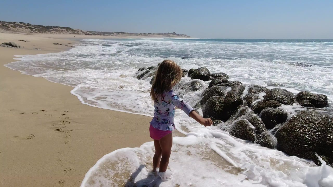 Young girl standing in foamy surf beside dark beach rocks.