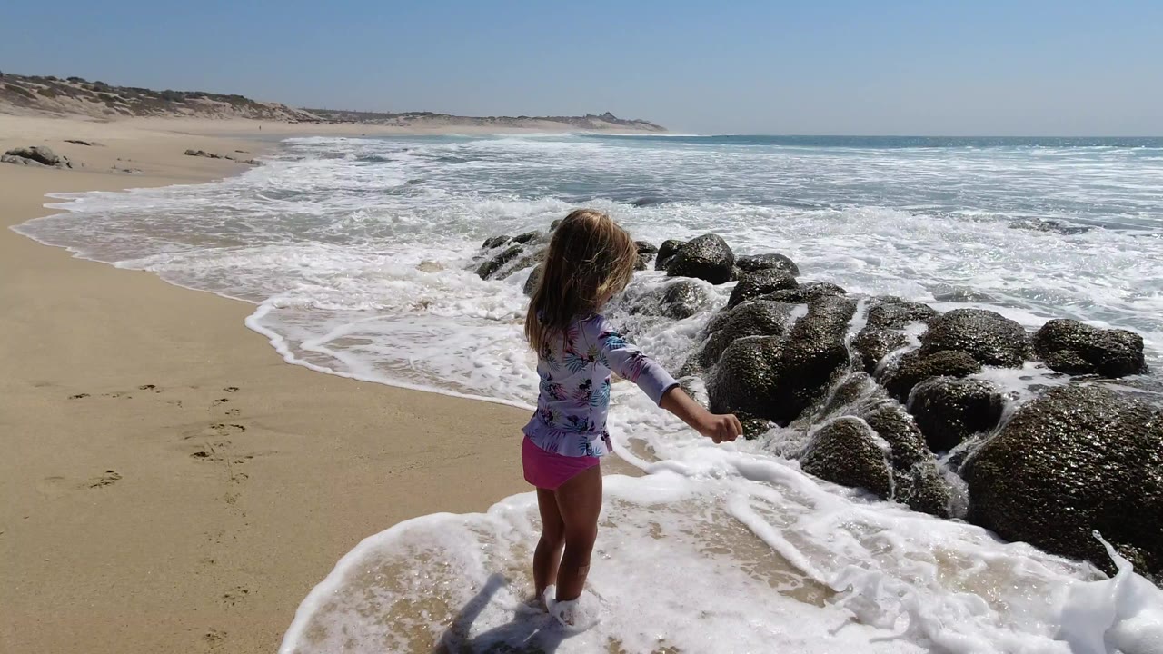The child remains near the waterline on the Los Cabos beach while white surf curls around her legs and the rock cluster fills the right side of frame.