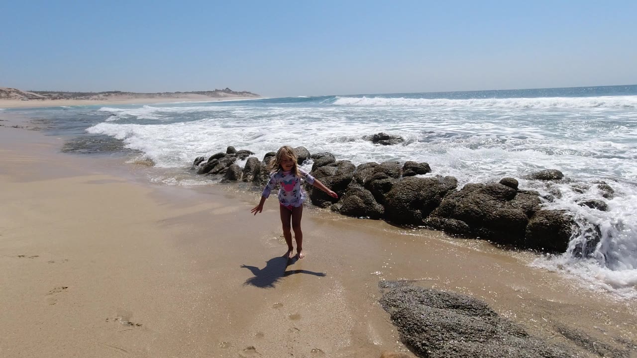 A child walks toward the camera on a sandy Los Cabos beach while white surf breaks around dark rocks behind her.