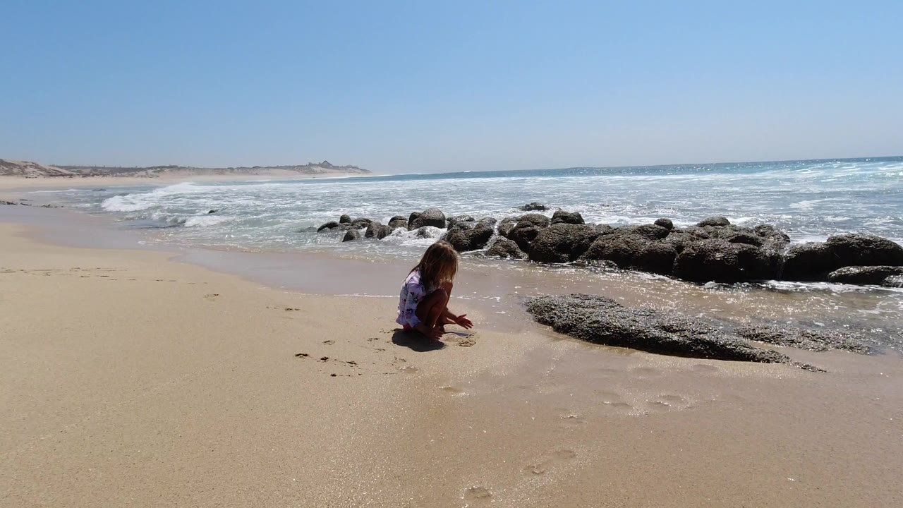 A child sits low on the sandy beach in Los Cabos, Mexico, near dark shoreline rocks as small waves break behind them under a clear sky.