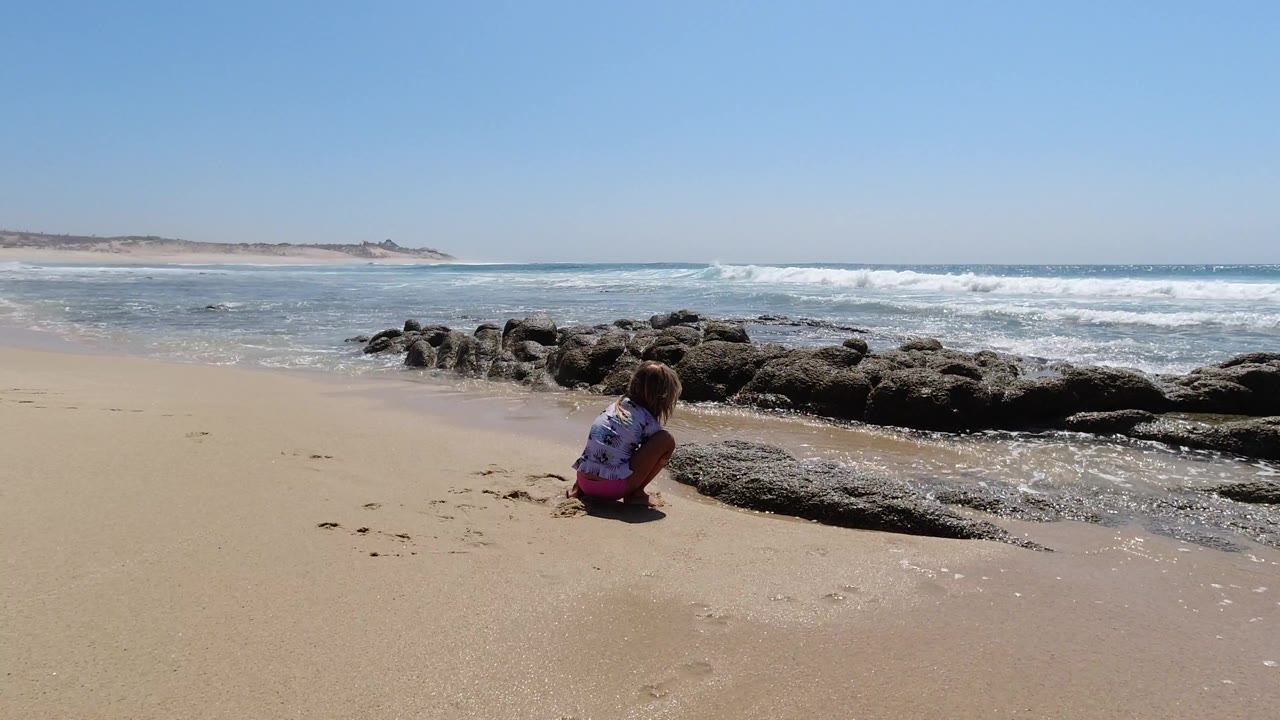At 0:01 in Los Cabos, Mexico, the girl remains crouched on the sand as a thin wash of water reaches the rock shelf in front of her.