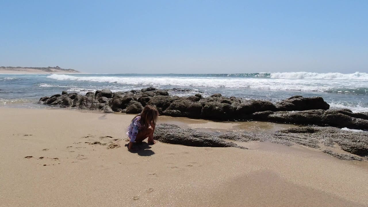 Child crouching in sand beside dark beach rocks and ocean surf.