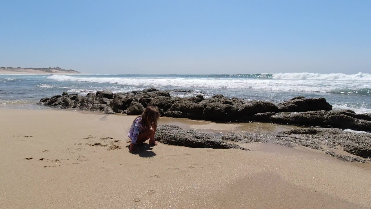 The child remains crouched near the sand and rocks as a line of white surf breaks across the Los Cabos shoreline in the background.