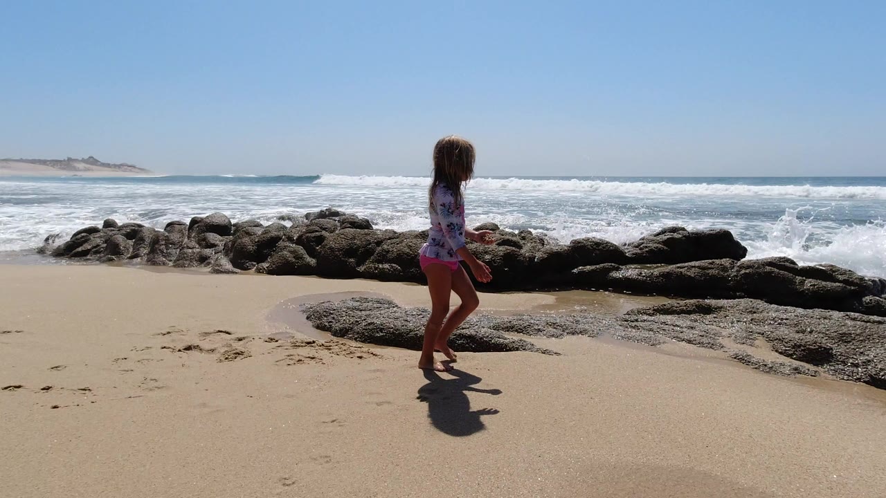 A young girl stands on a sandy Los Cabos beach near dark shoreline rocks, with white waves rolling across the ocean behind her.