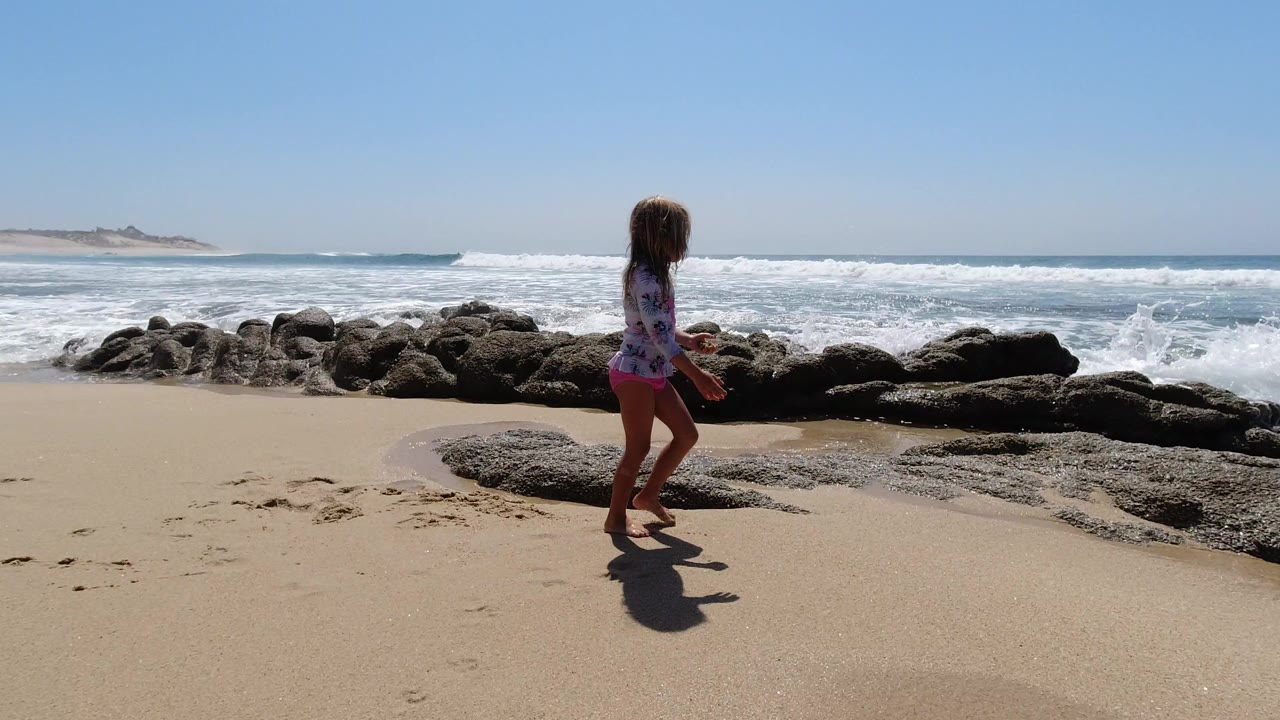 The girl moves across dry sand in Los Cabos as foamy surf breaks behind a line of low coastal rocks.