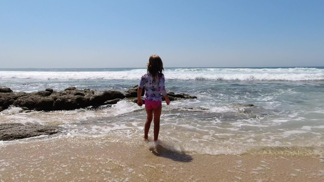 A young girl stands on wet sand in Los Cabos, Mexico, facing incoming waves with dark rocks to her left and a clear blue sky above.