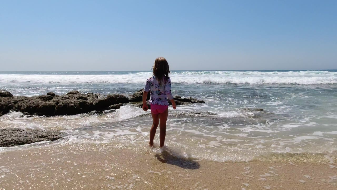 The child remains near center frame in Los Cabos, Mexico, watching white surf roll around low dark rocks under strong midday light.