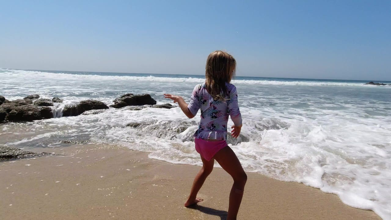 Young girl in a patterned swim shirt and pink bottoms stands near foamy surf on a Los Cabos beach, with dark rocks and a blue ocean horizon behind her.