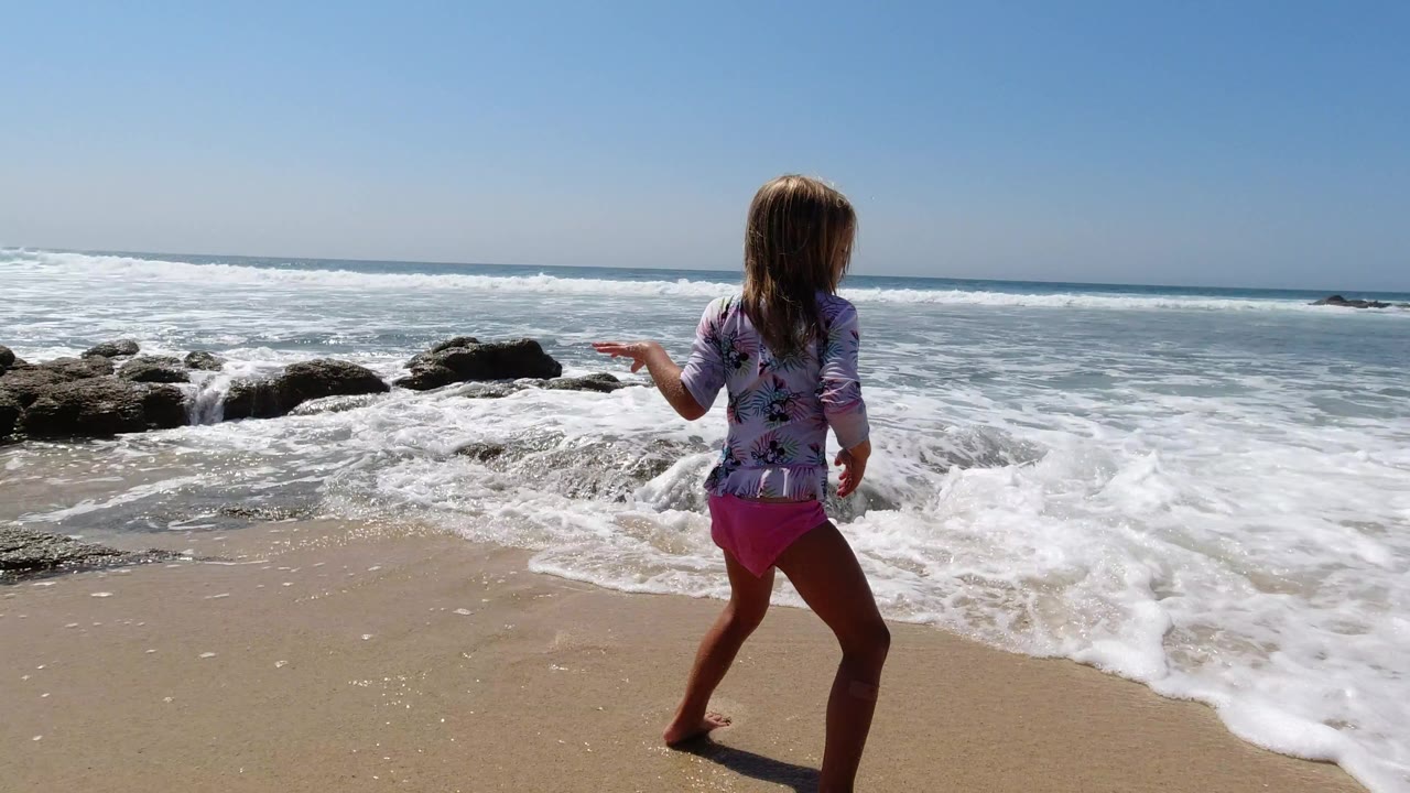 The young girl shifts her stance toward the incoming whitewater on the Los Cabos shoreline, with scattered rocks and a bright blue sky across the background.
