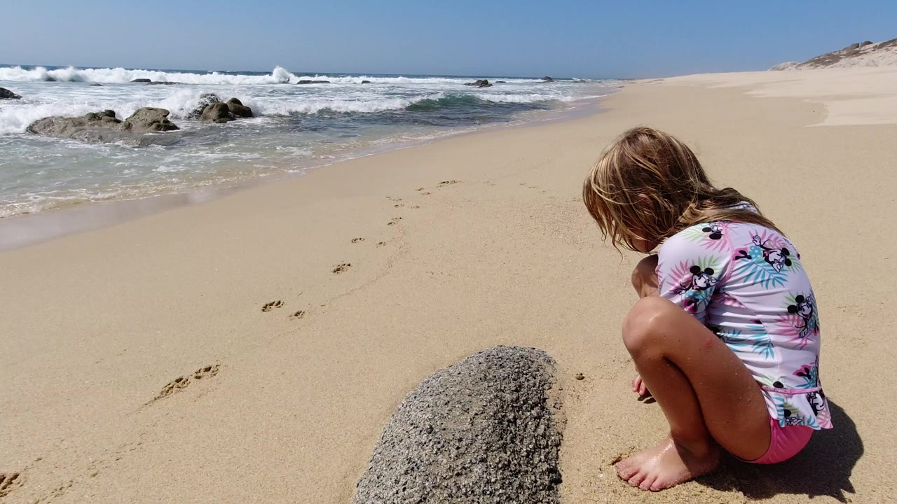 Girl crouching on sandy beach beside a rock and footprints near surf.
