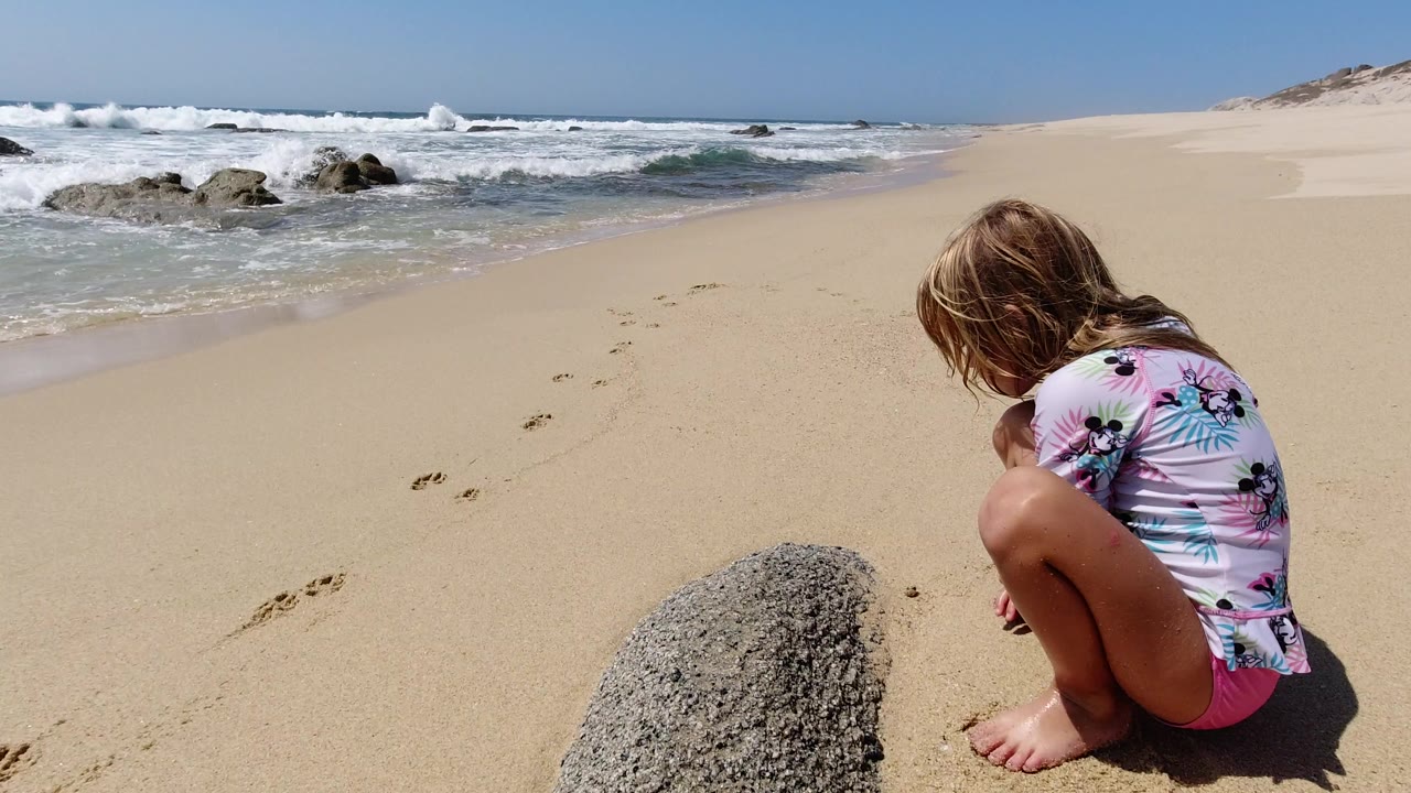 At 0:01 in Los Cabos, Mexico, the crouching girl keeps her head down toward the sand while white surf rolls behind her and footprints cross the open beach.