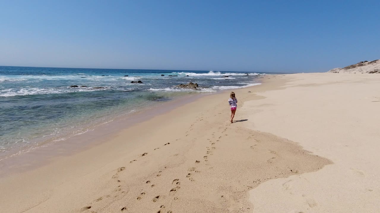Wide elevated view of a Los Cabos beach with a lone runner in pink shorts on pale sand, blue water at left, rocks offshore, and footprints leading toward the midground.