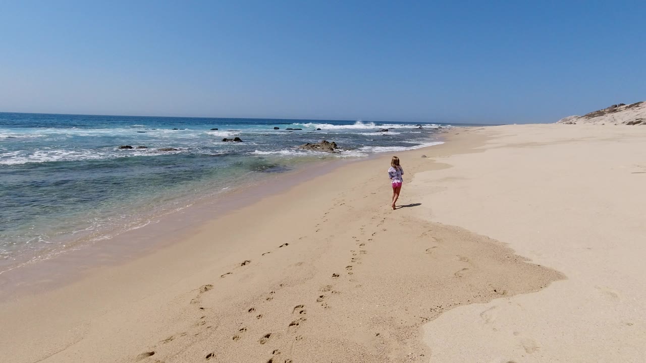 A lone runner continues along the Los Cabos shoreline, with shallow blue-green water and small breaking waves on the left and open sand stretching to dunes on the right.