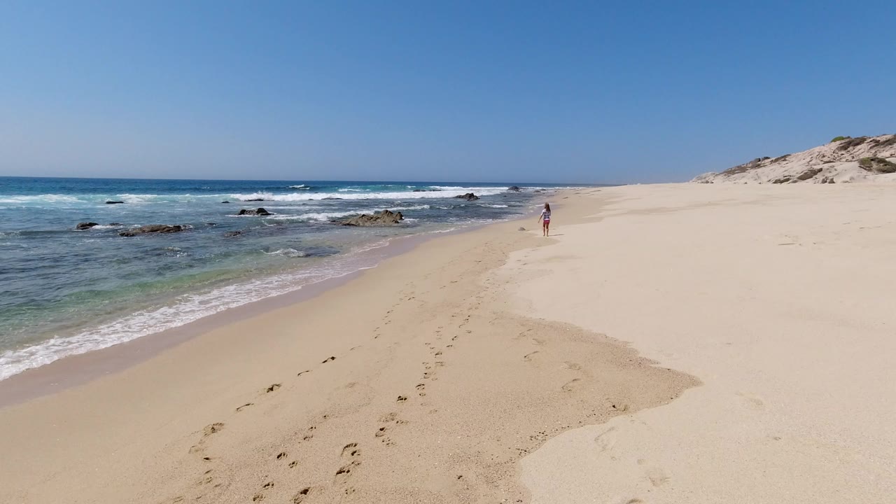 Los Cabos beach view at the start of the clip, with blue surf on the left, footprints in pale sand, dunes on the right, and a distant person near the shoreline.