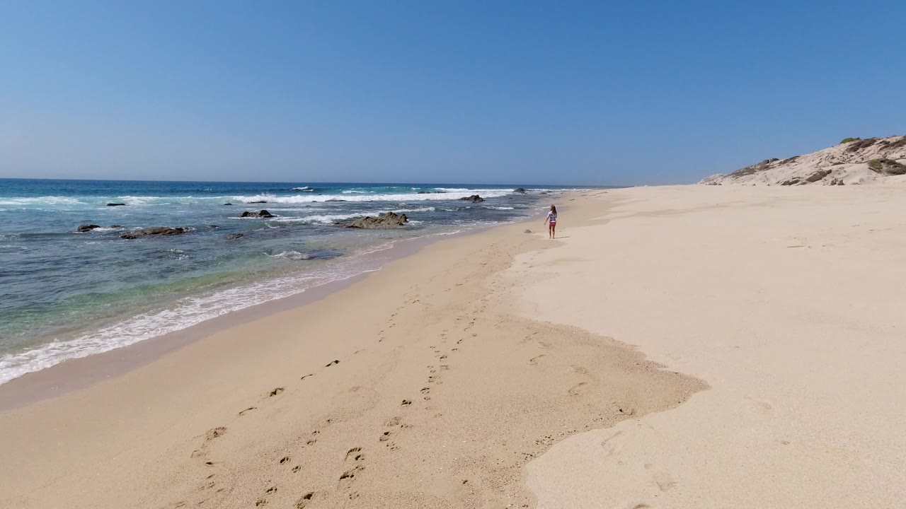 Los Cabos shoreline one second in, with low waves washing toward the sand, dark rocks offshore, a trail of footprints, and a small distant person ahead.