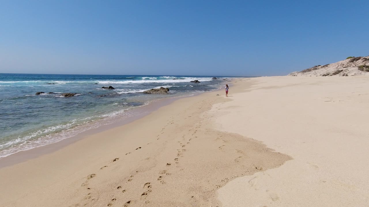 Wide coastal beach with turquoise water, surf rocks, sand, dunes, and a small distant person.