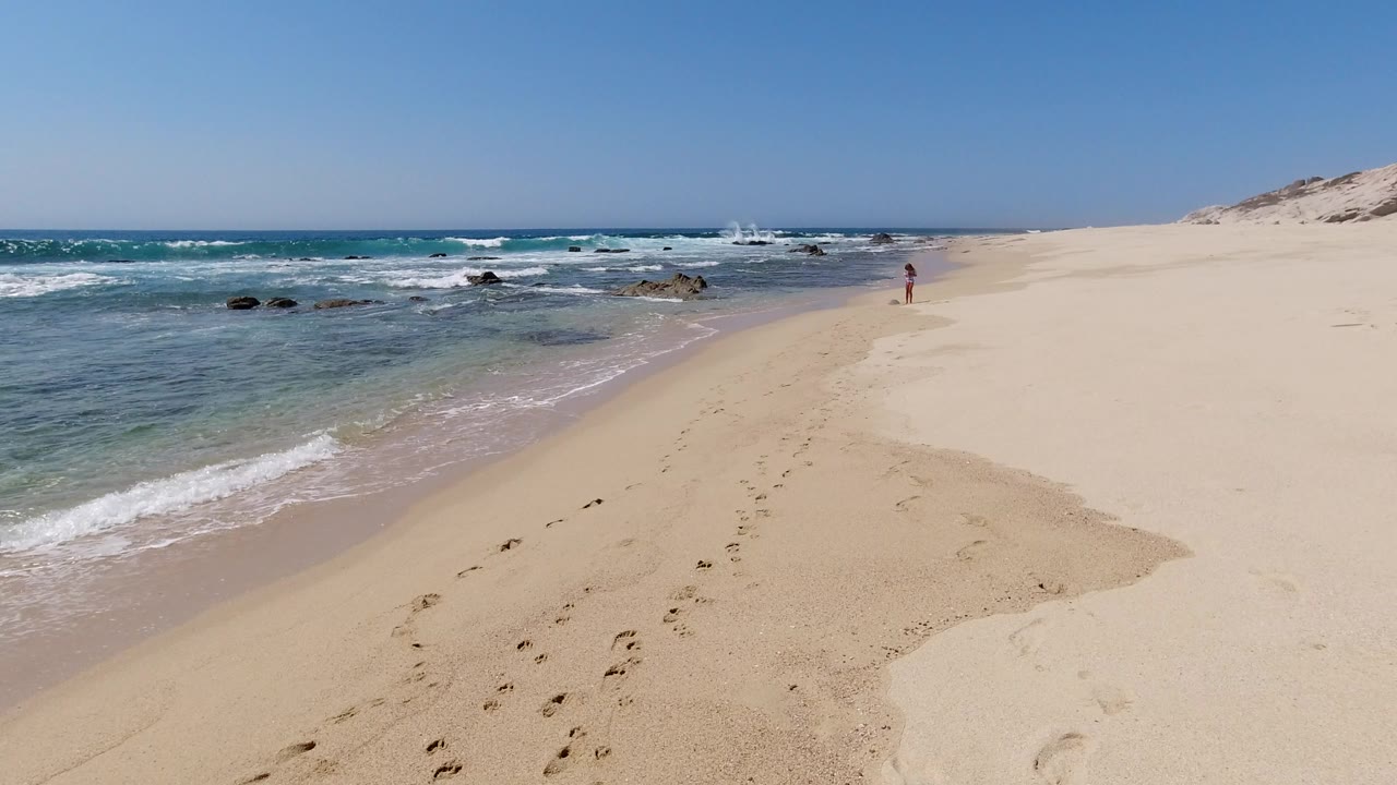 Wide sandy beach with surf, rocks, footprints, and a small child ahead.
