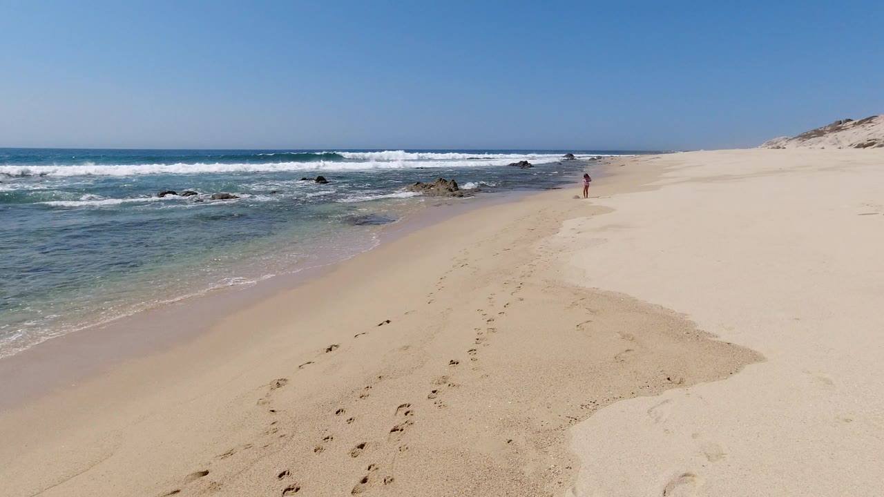 Wide sandy beach with surf, rocks, footprints, dunes, and a distant person.