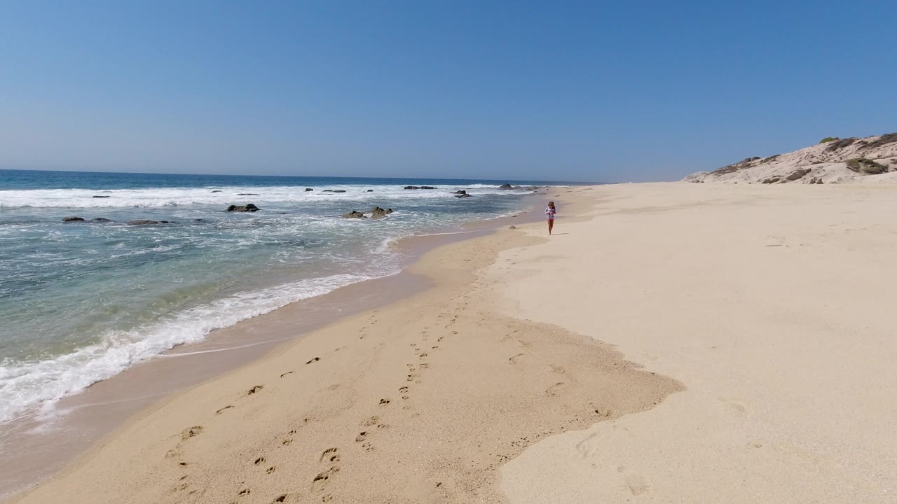 Los Cabos shoreline one second in, with the wave edge slightly farther up the sand, scattered dark rocks offshore, footprints in the foreground, and the distant walker near the right side.