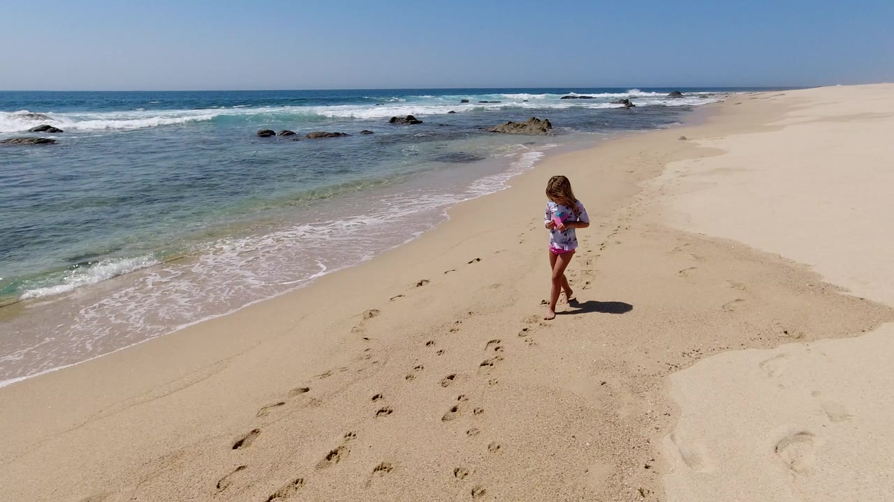 Young girl walking barefoot near footprints on a sandy beach beside ocean waves.