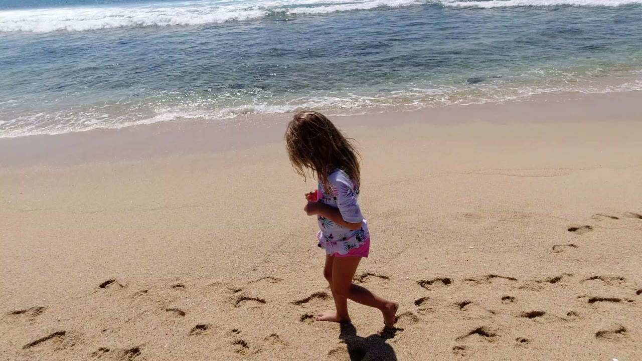 Young girl walking barefoot on sand beside ocean surf.