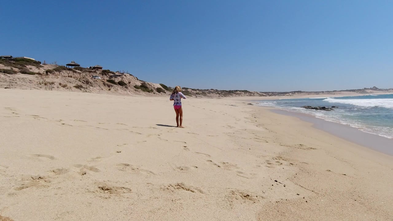 The person remains upright on the Los Cabos shoreline as small waves break to the right and the beach stretches into the distance under clear sky.