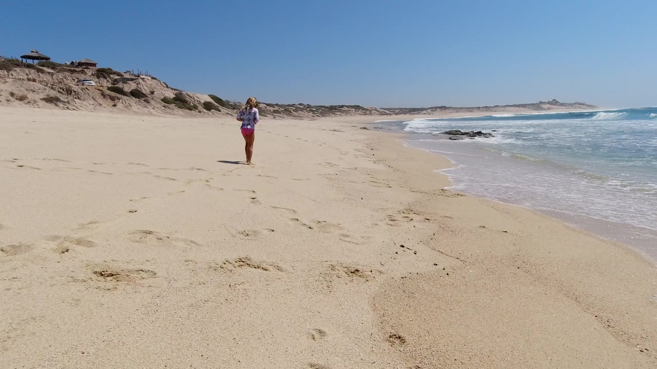 A young girl stands on a broad Los Cabos beach, with empty sand in front, low dunes and houses to the left, and blue surf breaking along the right edge.