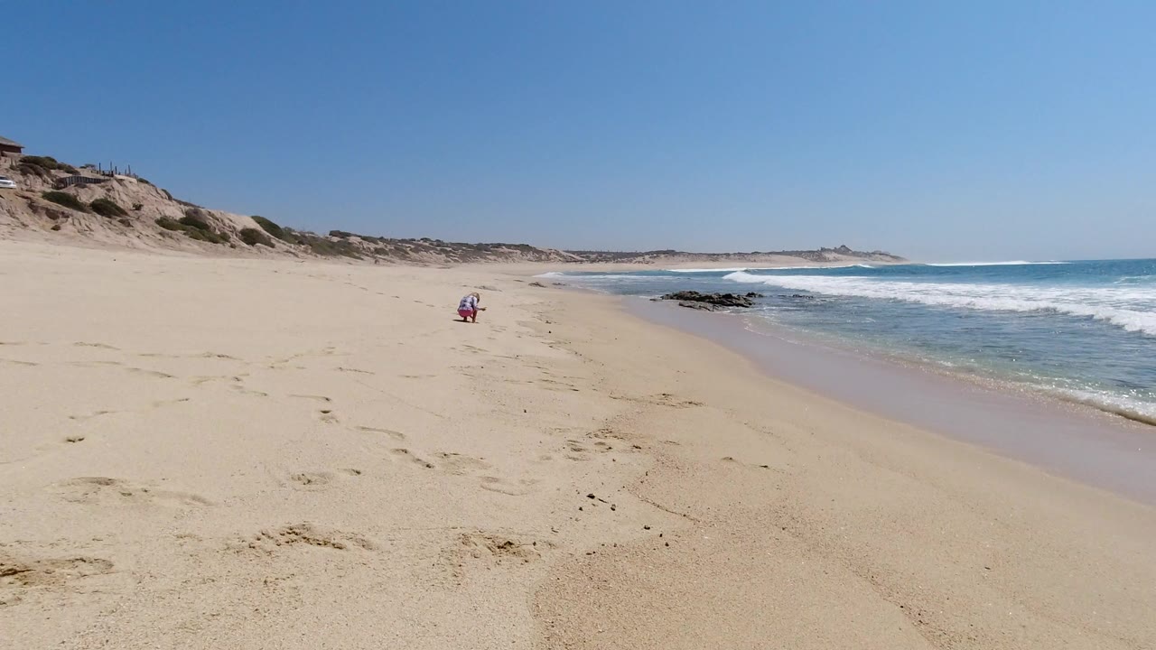 Opening view of a Los Cabos beach with pale sand in the foreground, low dunes to the left, white surf rolling on the right, and no visible people.